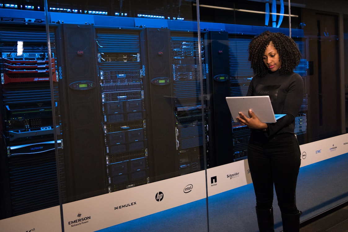 emale IT professional standing in front of a data center server rack, working on a laptop."
